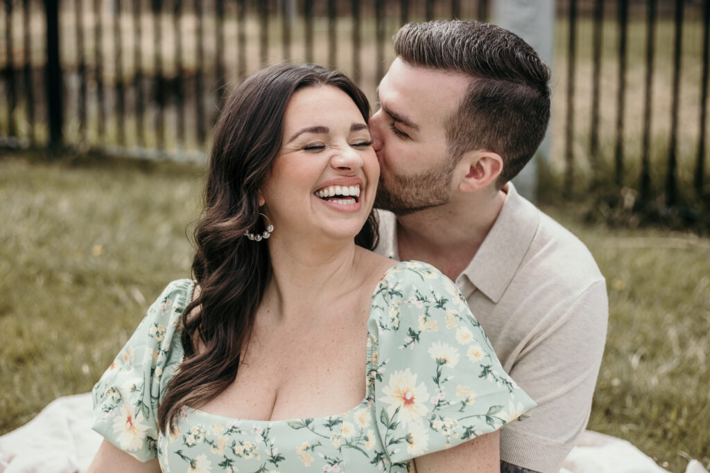 A brunette man whispering in the brunette woman's ear at the Paul Revere Heritage Park in Canton. Captured by Boston Engagement Photographer Hearts of Sage Photography